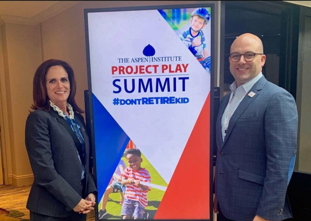 Stephanie and Brad Infante standing next to an Aspen Institute Project Play Summit sign